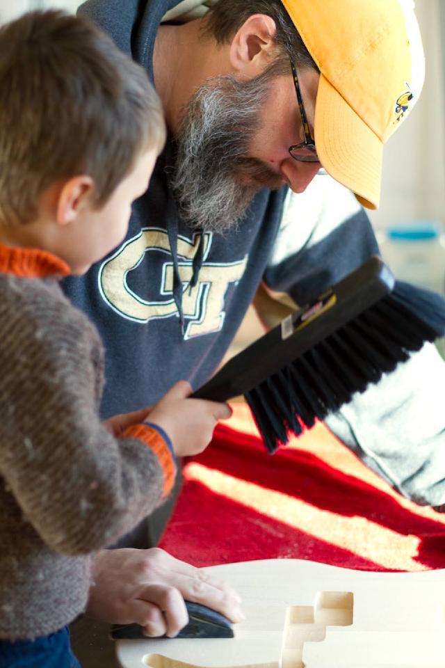 Bearded man sands guitar body while boy holding large brush looks on