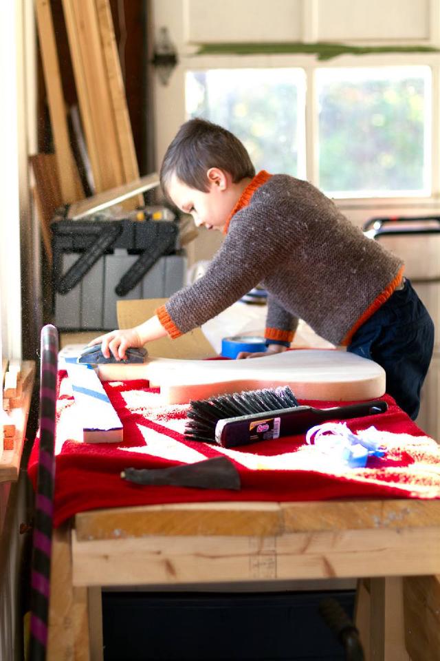 boy wearing a brown sweater and standing on a stool sands the body of a guitar