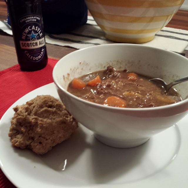 beef stew in a white bowl with a biscuit and a bottle of beer