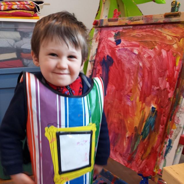 boy in a smock, beaming proudly, standing next to a painting