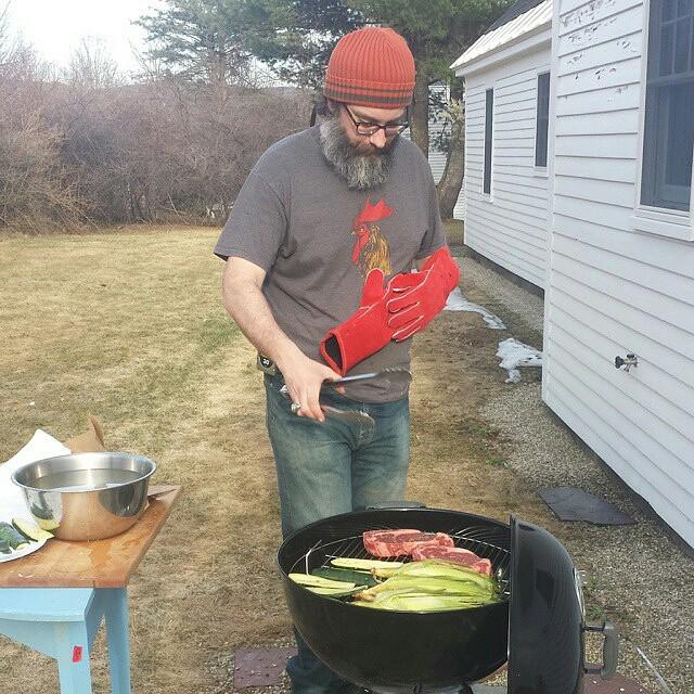 man with large beard, wearing orange beanie and blue jeans tends steaks and corn on a weber grill