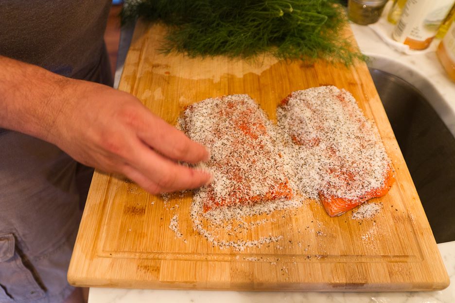 Hands sprinkle salt, sugar, and pepper mixture over salmon filets