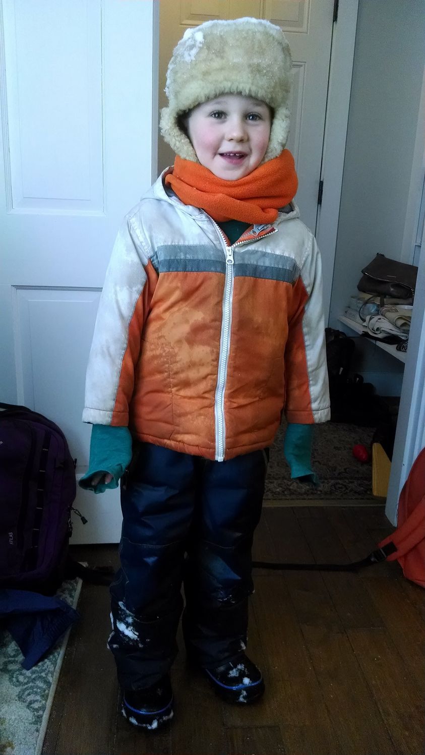 boy with cherry cheeks bundled up in hat, coat, and snow pants covered in snow